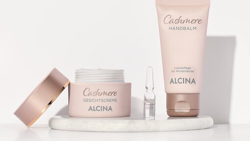 3 Produkte der Hautpflege-Serie mit Cashmere stehen auf einem Tablett. Neben einem geöffneten Cremetiegel in beiger Farbe sieht man mittig eine gläserne Ampulle sowie eine beige auf dem Kopf stehende Tube Handcreme