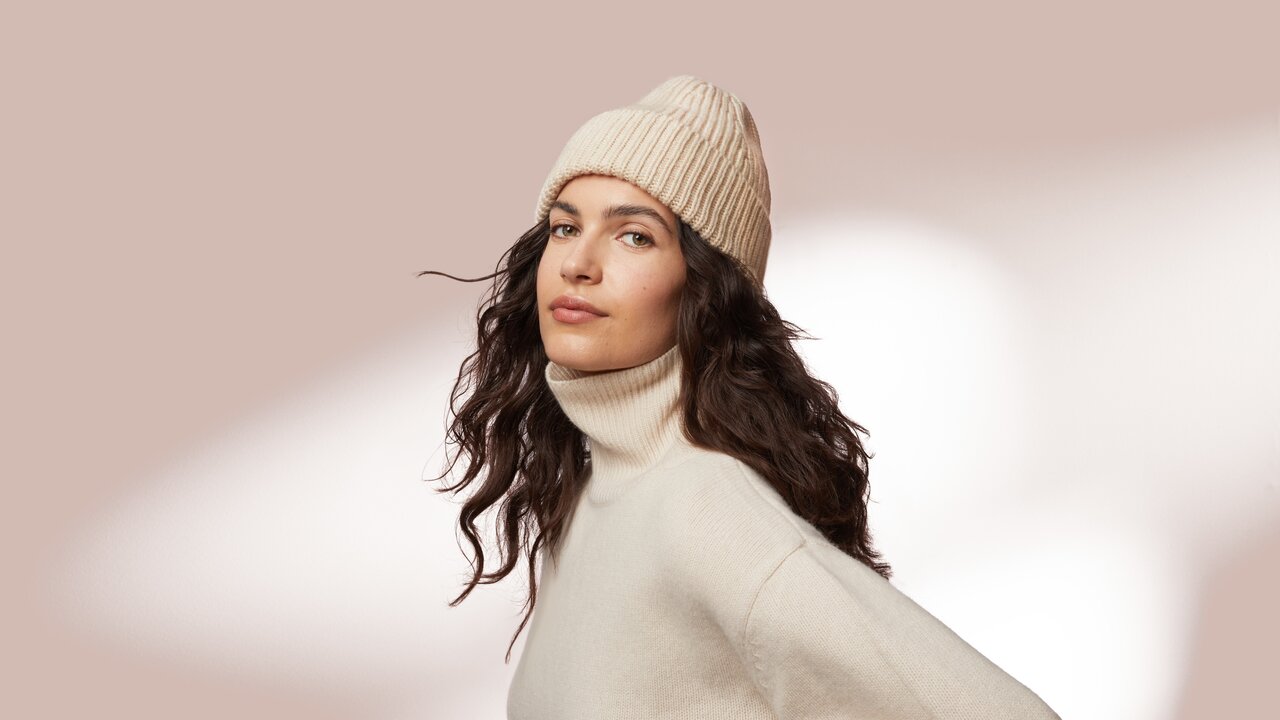 So Schützen Sie Ihre Haut gegen die Kälte! Portrait einer jungen Frau mit schöner Haut und langen lockigen Haaren, die einen Rollkragen Pullover und eine Wintermütze trägt auf rosa Hintergrund als Sinnbild für Gesichtspflege bei trockener Haut im Winter
