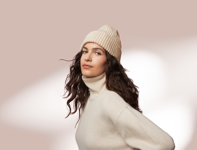 Portrait einer jungen Frau mit schöner Haut und langen lockigen Haaren, die einen Rollkragen Pullover und eine Wintermütze trägt auf rosa Hintergrund als Sinnbild für Gesichtspflege bei trockener Haut im Winter 