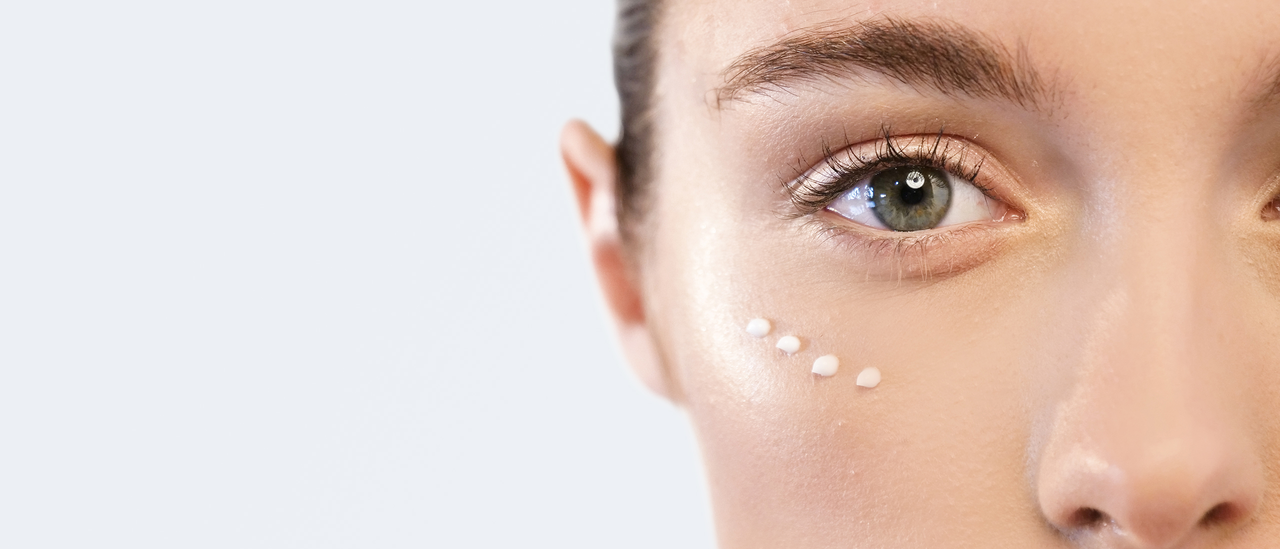 Nahaufnahme vom Gesicht einer schönen Frau, die vier Tropfen einer Augencreme unter dem Auge hat als Sinnbild für die richtige Augenpflege bei Augenringen und Augenfalten