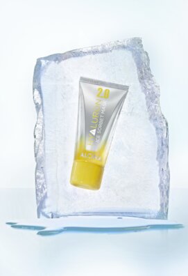 Eine Tube der Alcina Hyaluron 2.0 Ice Sorbet Mask, eingefasst in einem klaren Eisblock, mit sichtbaren Wassertropfen am Boden.