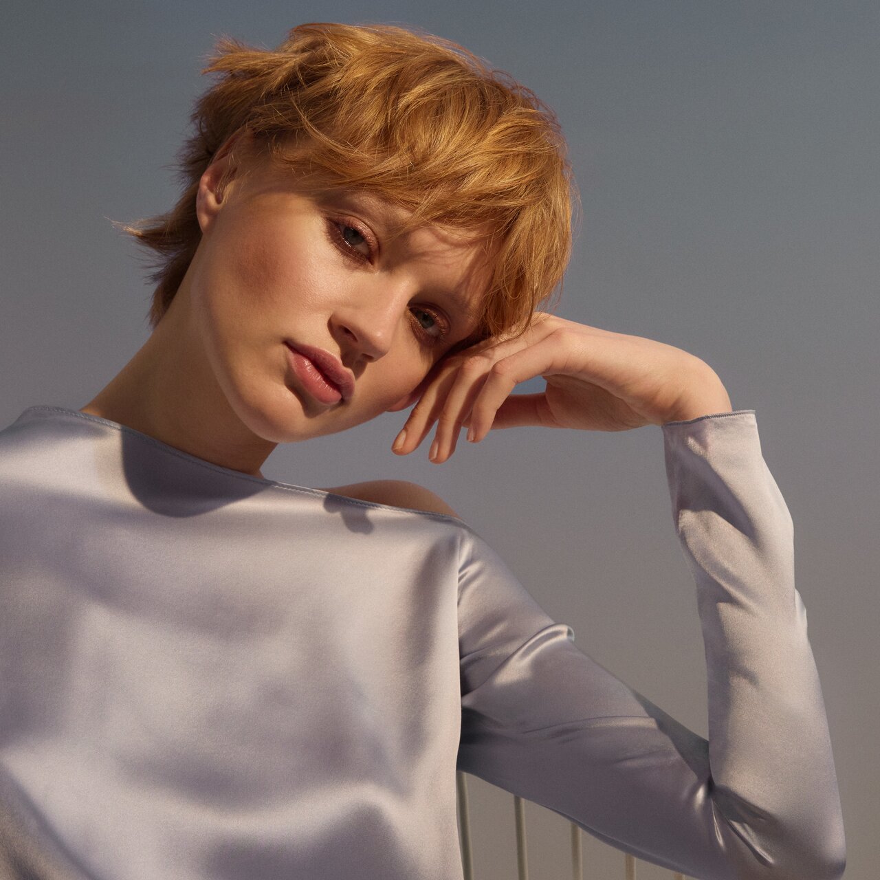 Porträt einer jungen Frau mit honigblonden Haaren vor hellblauem Hintergrund, die nachdenklich zum Betrachter blickt