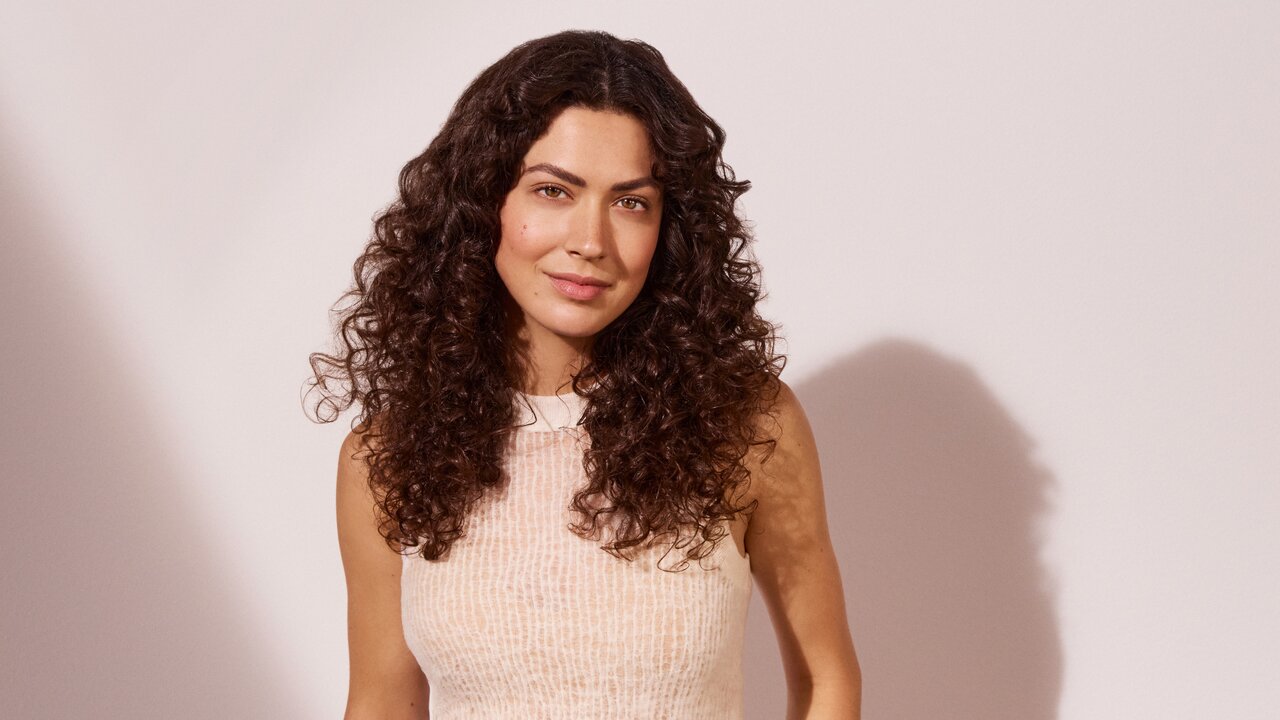  Frau mit langen dunkelbraunen Locken, die ein beigefarbenes Satinhemd trägt, vor einer hellen, rosefarbenen Wand. 