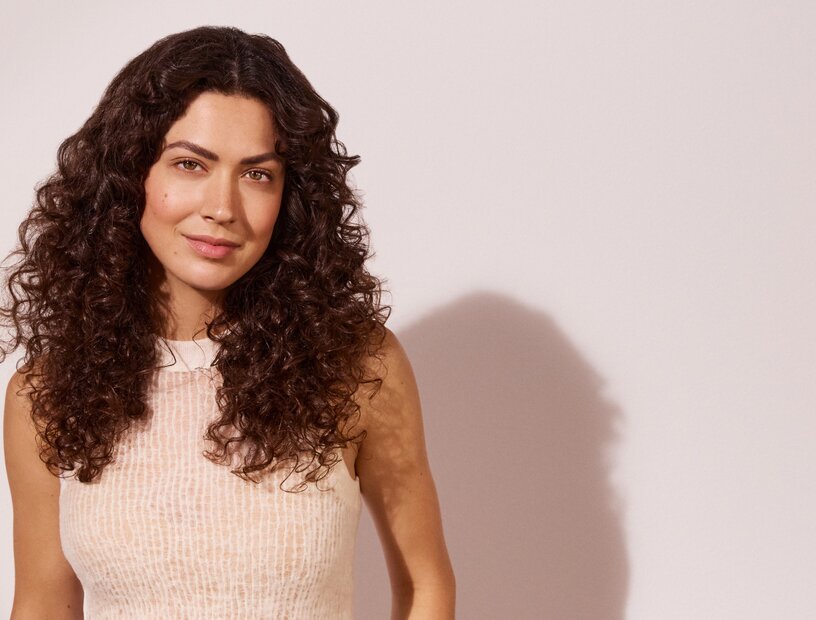  Frau mit langen dunkelbraunen Locken, die ein beigefarbenes Satinhemd trägt, vor einer hellen, rosefarbenen Wand. 