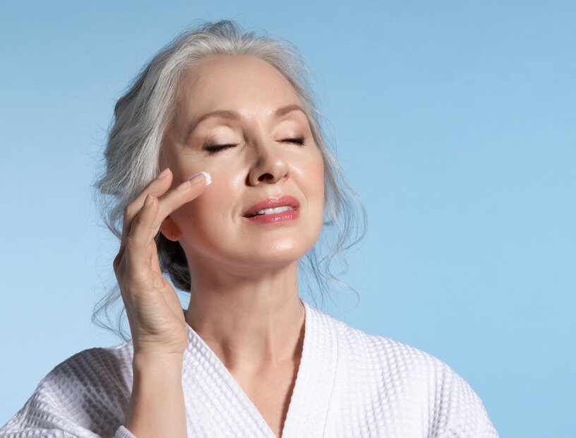Frau mit geschlossenen Augen und Gesicht nach oben gewandt, tupft mit der Fingerspitze einer Hand Gesichtspflege auf die glatte Wange