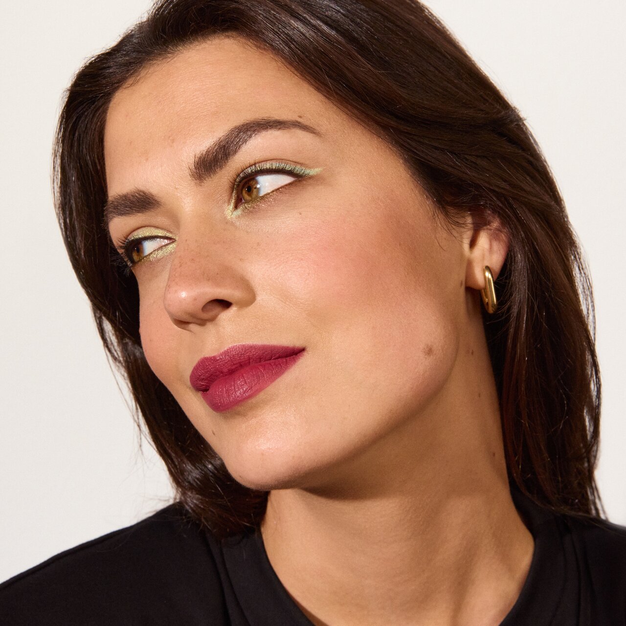 Schöne junge Frau mit schulterlangen dunklen Haaren schaut lächelnd über ihre Schulter nach hinten. Sie trägt einen zartgrünen Lidstrich und matten beerenfarbigen Lippenstift.