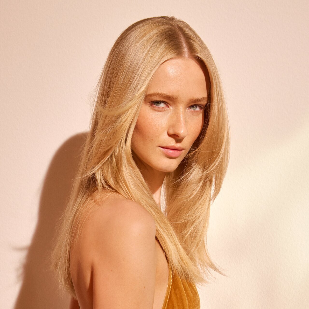 Blonde junge Frau mit langen Haaren schaut lächelnd seitwärts über ihre Schulter in Richtung Betrachter. 