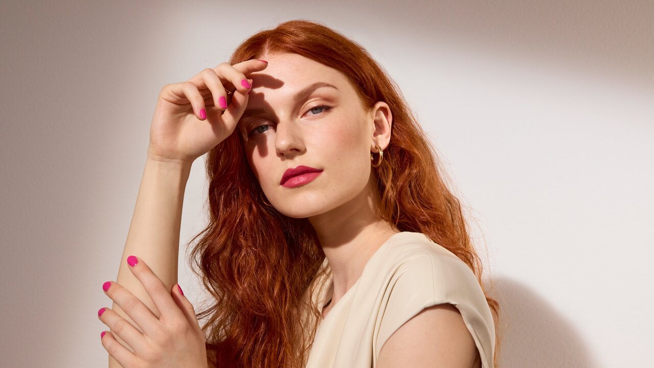 Schöne Frau mit langen roten Haaren präsentiert die Make-up-Trends im Frühjahr und Sommer 2026, ihre Fingernägel sind pink lackiert, ihre Lippen sind roséfarben geschminkt, auf den Wangen und Augen trägt sie schimmernden Bronzer und Highlighter 