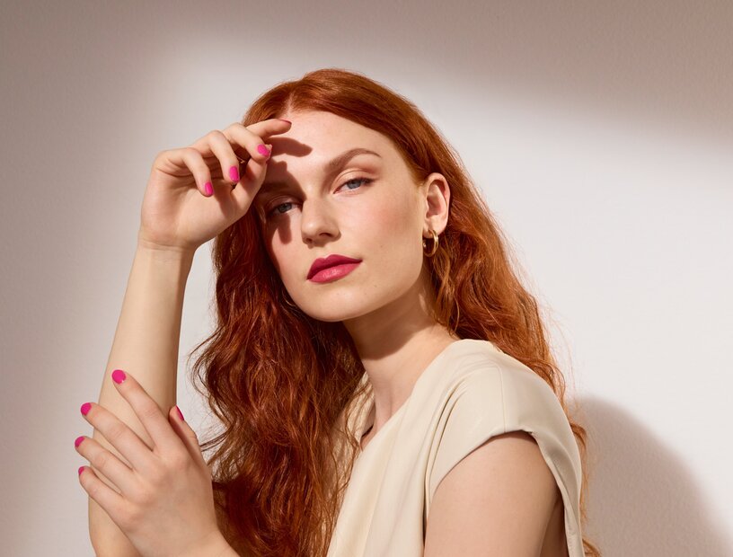 Schöne Frau mit langen roten Haaren präsentiert die Make-up-Trends im Frühjahr und Sommer 2026, ihre Fingernägel sind pink lackiert, ihre Lippen sind roséfarben geschminkt, auf den Wangen und Augen trägt sie schimmernden Bronzer und Highlighter 
