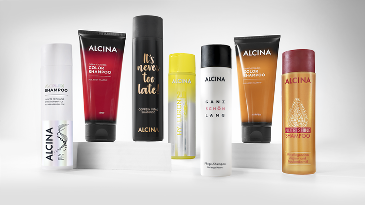 Sieben ALCINA Shampoos in unterschiedlich gestalteten Flaschen und Tuben stehen nebeneinander auf weißen Podesten.