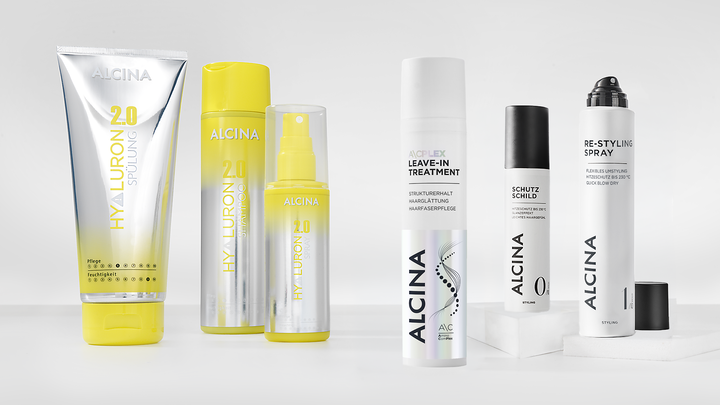 Sechs ALCINA Haarpflegeprodukte zum Hitzeschutz, darunter eine Tube, Sprays und ein Leave-in-Treatment, stehen auf weißen Podesten.