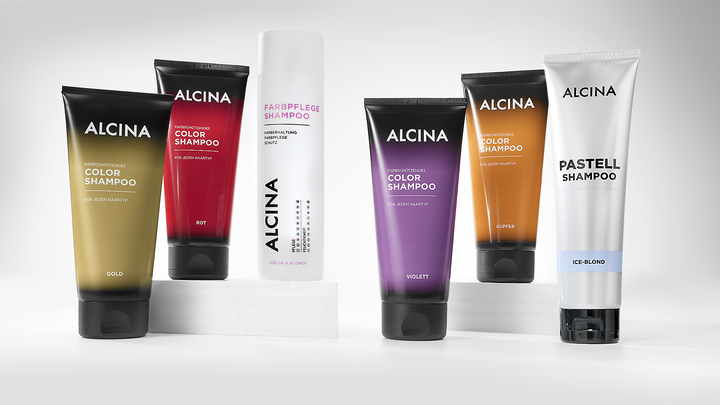 Sechs ALCINA Farbshampoos in goldenen, roten, weißen, violetten und kupferfarbenen Verpackungen stehen auf weißen Podesten.