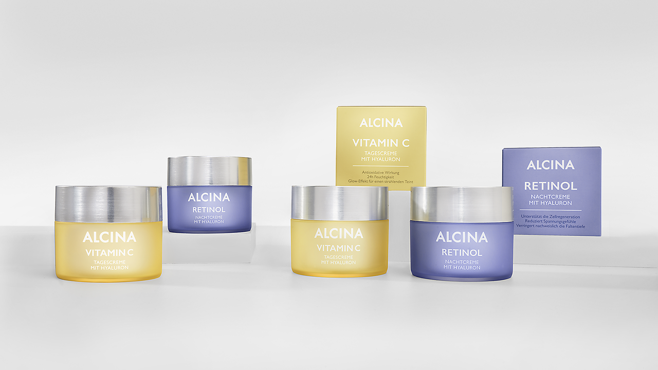 Glow-Effekt am Tag und Regeneration in der Nacht – die ALCINA Vitamin-Serie Je zwei Tiegel ALCINA Vitamin C Creme mit gelber Verpackung und Tiegel ALCINA Retinol Creme mit violetter Verpackung auf durchsichtigem Sockel platziert