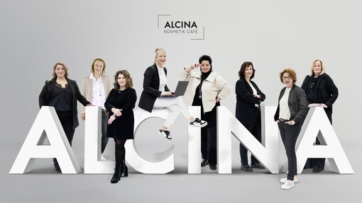 ALCINA Kosmetik Cafe  Acht ALCINA Beauty Expertinnen des Kosmetik Café sitzen oder stehen auf einem übergroßen, dreidimensionalem ALCINA Logo