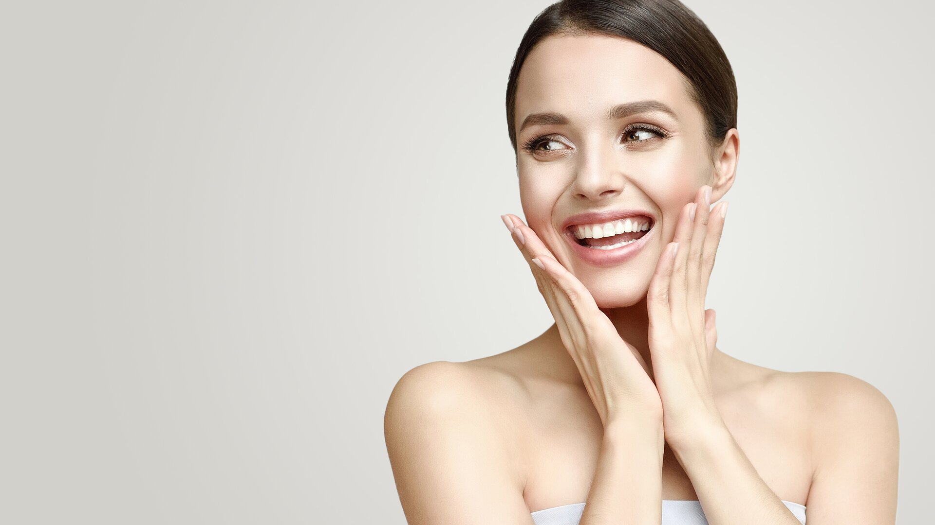 Hautpflege für schöne Haut braucht passende Produkte für die Gesichtspflege und -reinigung, Augen- und Lippenpflege und für die Hand- und Körperpflege. Lächelnde Frau mit schöner, gepflegter Haut, die zur Seite blickt und die Hände jeweils links und rechts an die Wangen legt