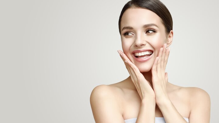 Hautpflege für schöne Haut braucht passende Produkte für die Gesichtspflege und -reinigung, Augen- und Lippenpflege und für die Hand- und Körperpflege. Lächelnde Frau mit schöner, gepflegter Haut, die zur Seite blickt und die Hände jeweils links und rechts an die Wangen legt