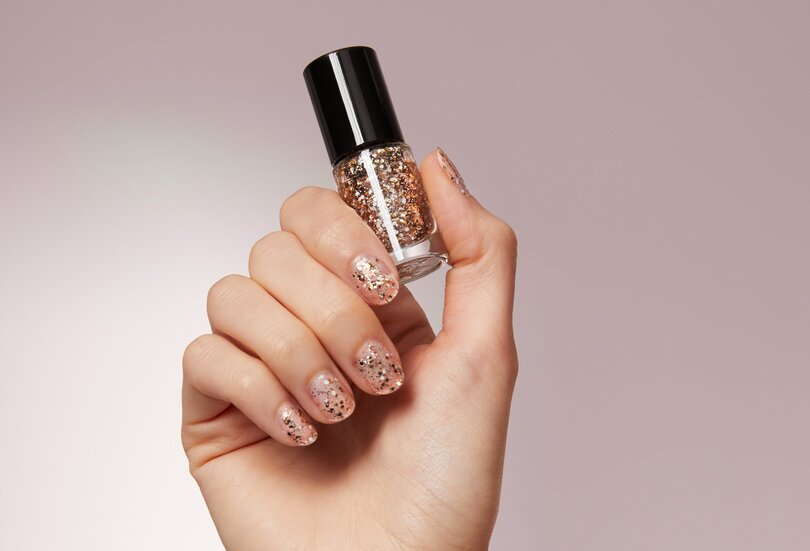 Besonders Highlight für den festlichen Look: Der “Golden Star”-Nagellack kann sowohl alleine als auch über dem Nagellack getragen werden Nahaufnahme einer gepflegten Hand und lackierten Nägeln mit Glitzer-Partikeln mit dem Trend-Nagellack Golden Star als Make-up-Trend Smoked-out Wings im Herbst Winter 2023