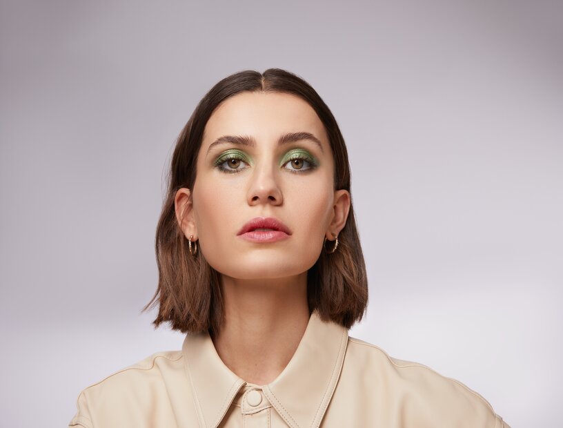 Portrait einer schönen jungen Frau mit kurzen braunen Haaren und stark geschminkten Augen mit grünem Lidschatten und Metallic Effekt als Sinnbild für die Make-up-Trends im Herbst Winter 2023